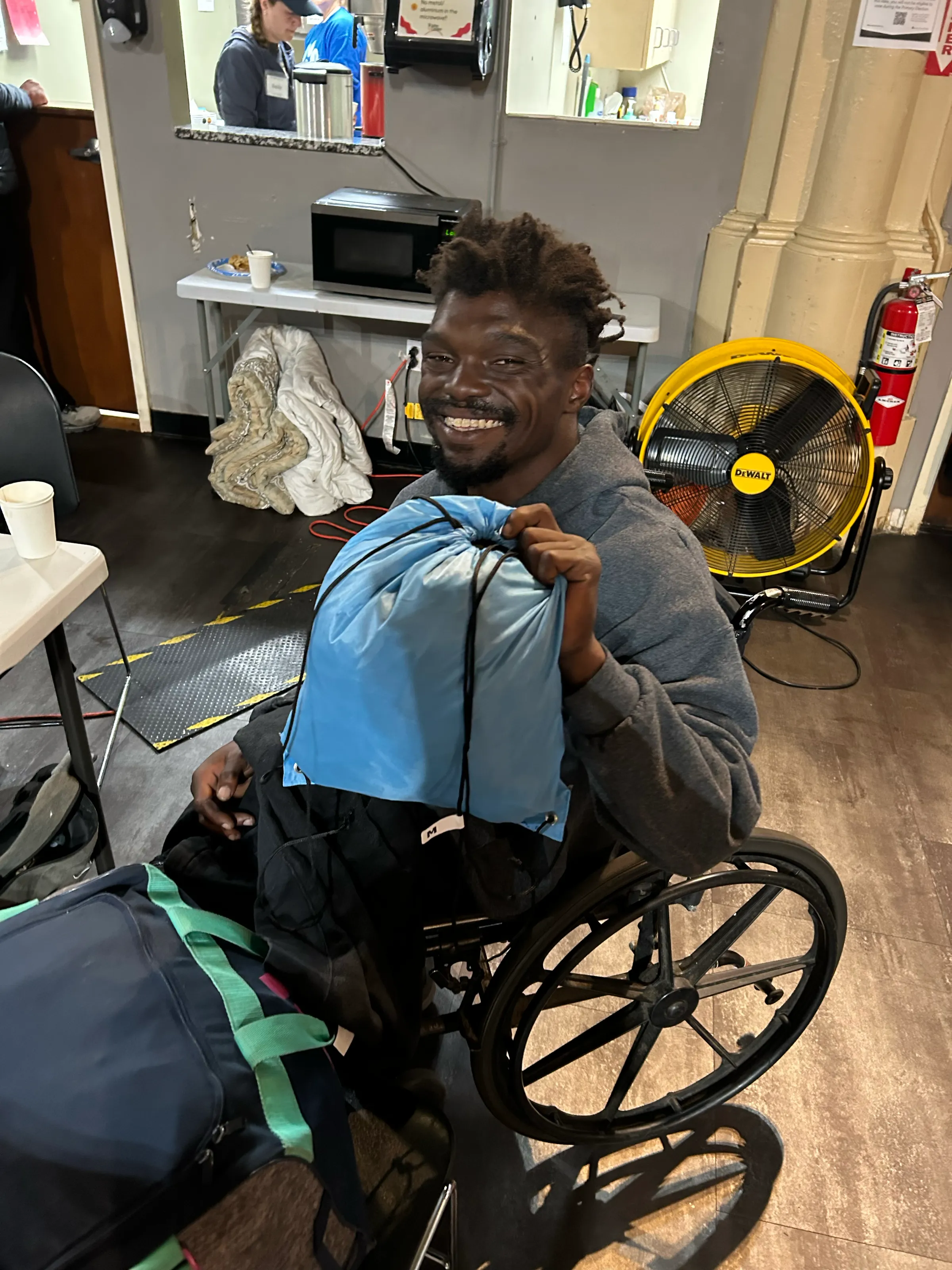 A man in a wheelchair smiles as he holds his blue Touch of Terra backpack at a shelter.